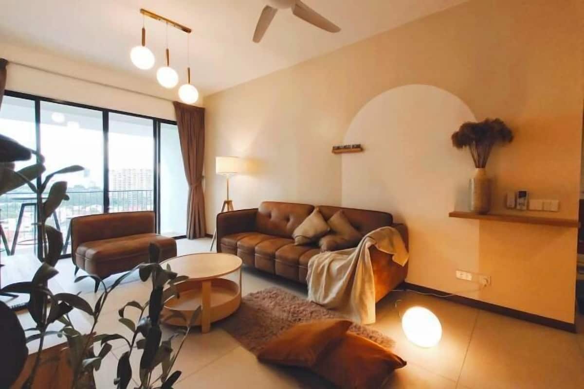 A modern living room with a sofa, armchair, and coffee table. There is a large window with curtains, a floor lamp, and a vase with dried flowers on a shelf.