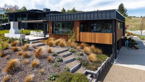 A modern house with a large deck, a stone pathway, and a landscaped garden with grasses and shrubs. - Modern 3 Bedroom Home in Wanaka with Mountain View (Wanaka)