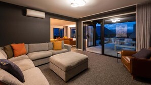 A modern living room with a sectional sofa, an ottoman, and a leather armchair. There is a glass door leading to a balcony with outdoor seating. - Modern 3 Bedroom Home in Wanaka with Mountain View (Wanaka)