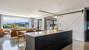 A modern kitchen with a large island, a dining area with chairs and a sofa, and a view of a mountain through large windows. - Modern 3 Bedroom Home in Wanaka with Mountain View (Wanaka)