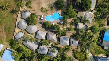 An aerial view of a resort with a swimming pool, thatched-roof structures, and palm trees.