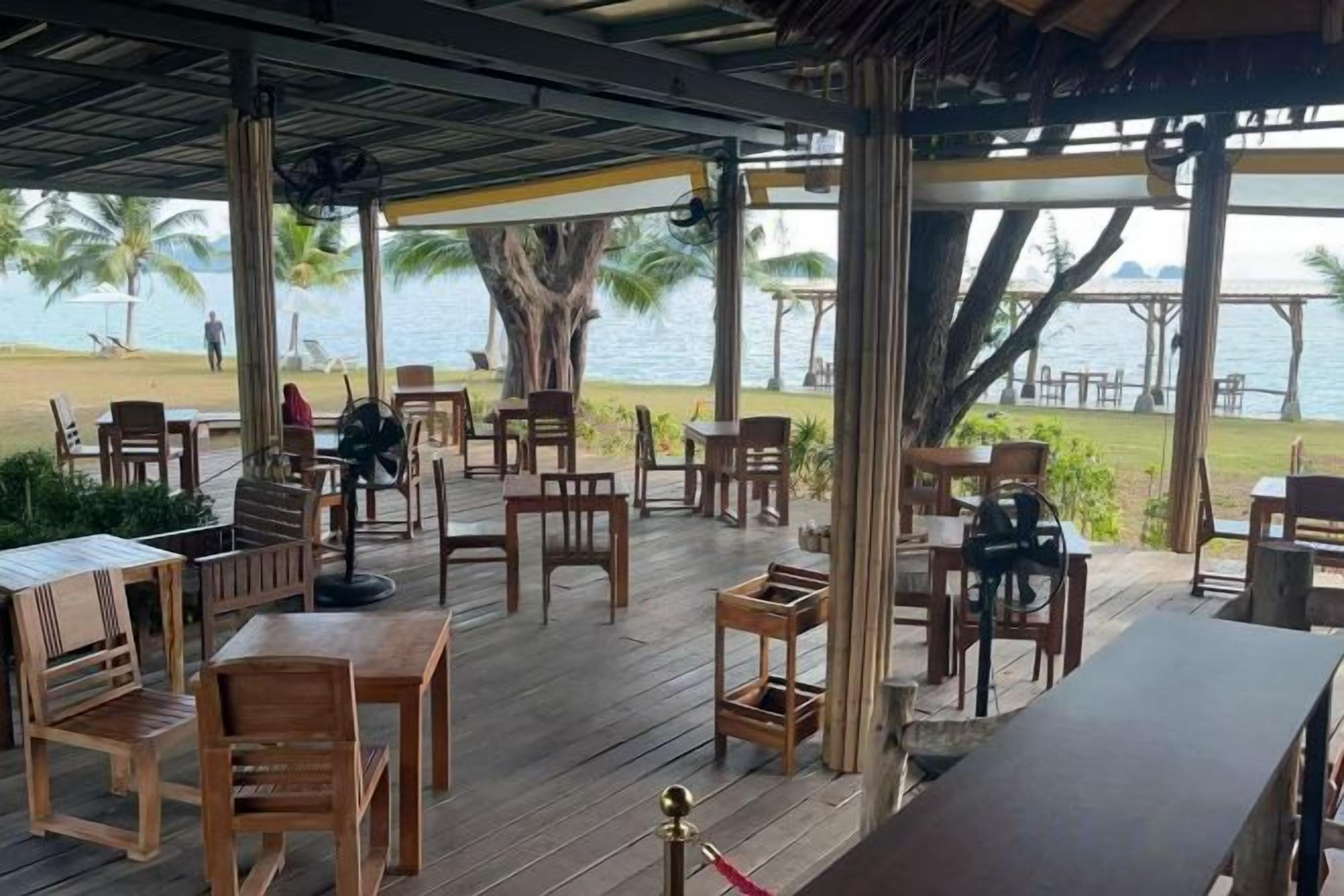 An outdoor seating area with wooden tables and chairs, overlooking a beach and palm trees.