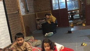 Een kamer met een bakstenen muur, een houten deur en een raam. Twee kinderen zitten op de vloer, waarvan er een een boek leest en de andere speelt. Een derde kind zit op een stoel op de achtergrond.