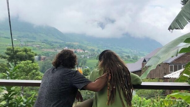 Een koppel zit op een balkon met uitzicht op een bergachtig landschap, met huizen in de verte.