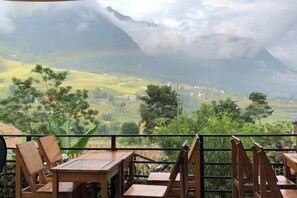 A table and chairs on a balcony overlooking a valley with a mountain and a rainbow.