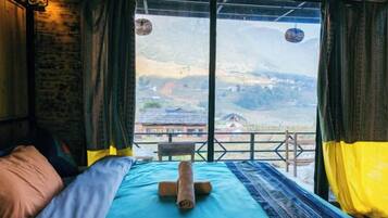 A room with a bed, a view of a mountainous landscape, and a balcony with a chair.