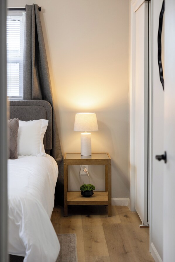 Un dormitorio con cama, una mesita de noche con lámpara, una planta pequeña y una puerta.