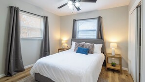 Un dormitorio con una cama, dos mesitas de noche, un ventilador de techo y una ventana con cortinas.