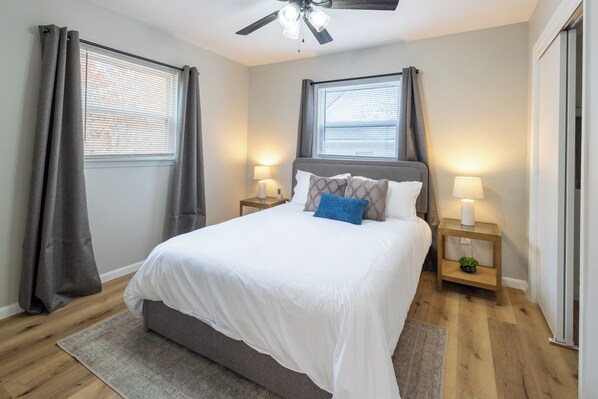 Une chambre à coucher comprenant un lit, deux tables de chevet, un ventilateur de plafond et une fenêtre avec des rideaux.