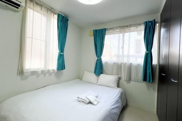 A neatly made bed with white linens and a folded towel, a window with teal curtains, and a dark-colored wardrobe. - JING HOUSE ShinTakasgo (Tokyo)