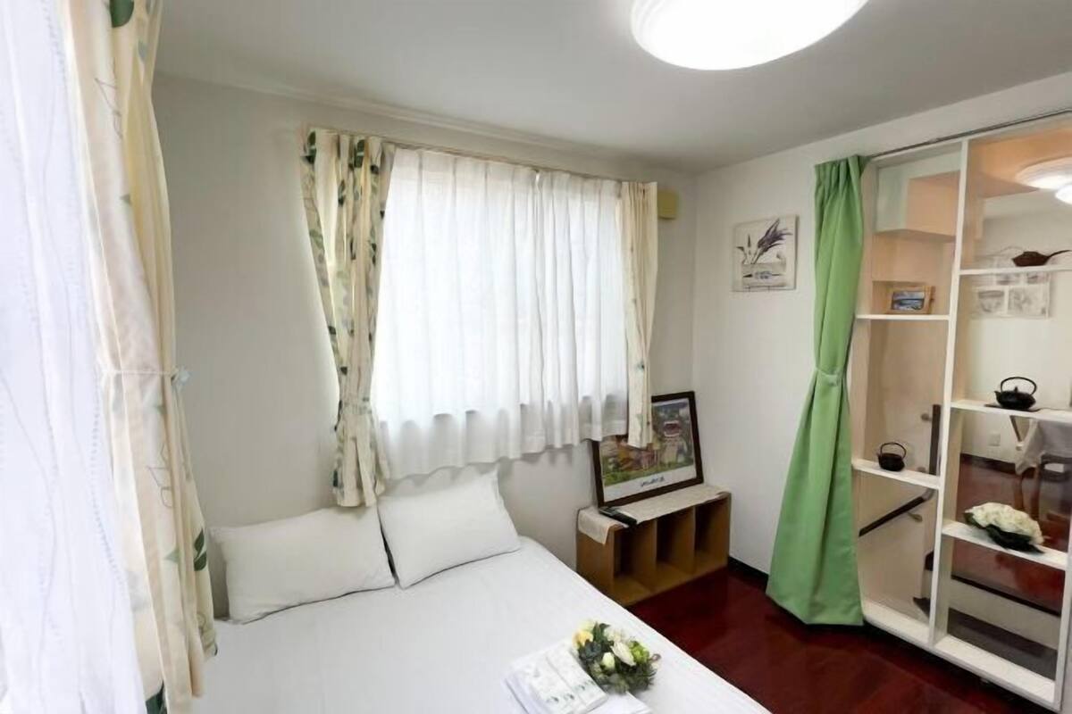 A hotel room with a bed, a bedside table, a framed picture, a green curtain, and a white shelf with items.