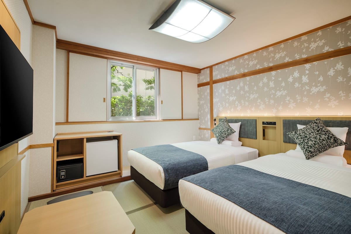 A hotel room with two beds, a TV, a desk, and a window with a view of greenery.