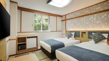 A hotel room with two beds, a TV, a desk, and a window with a view of greenery.