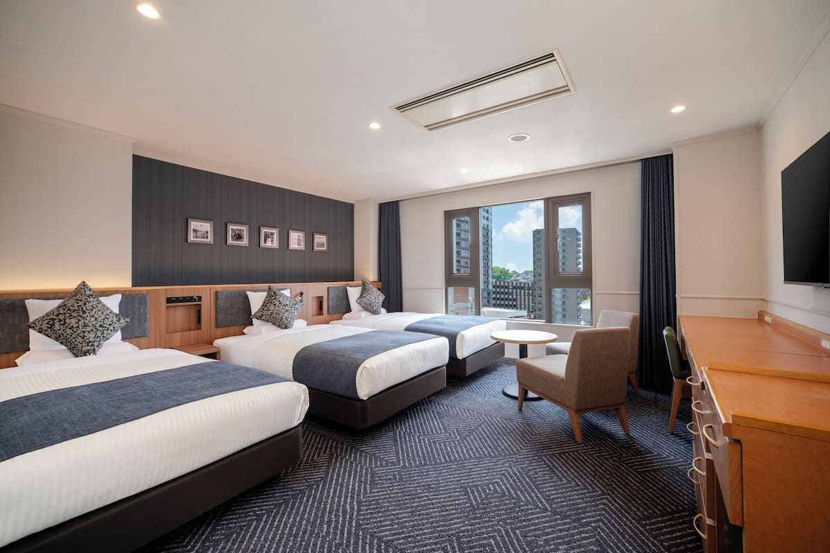 A hotel room with three beds, a desk, a chair, a TV, and a window with a city view.