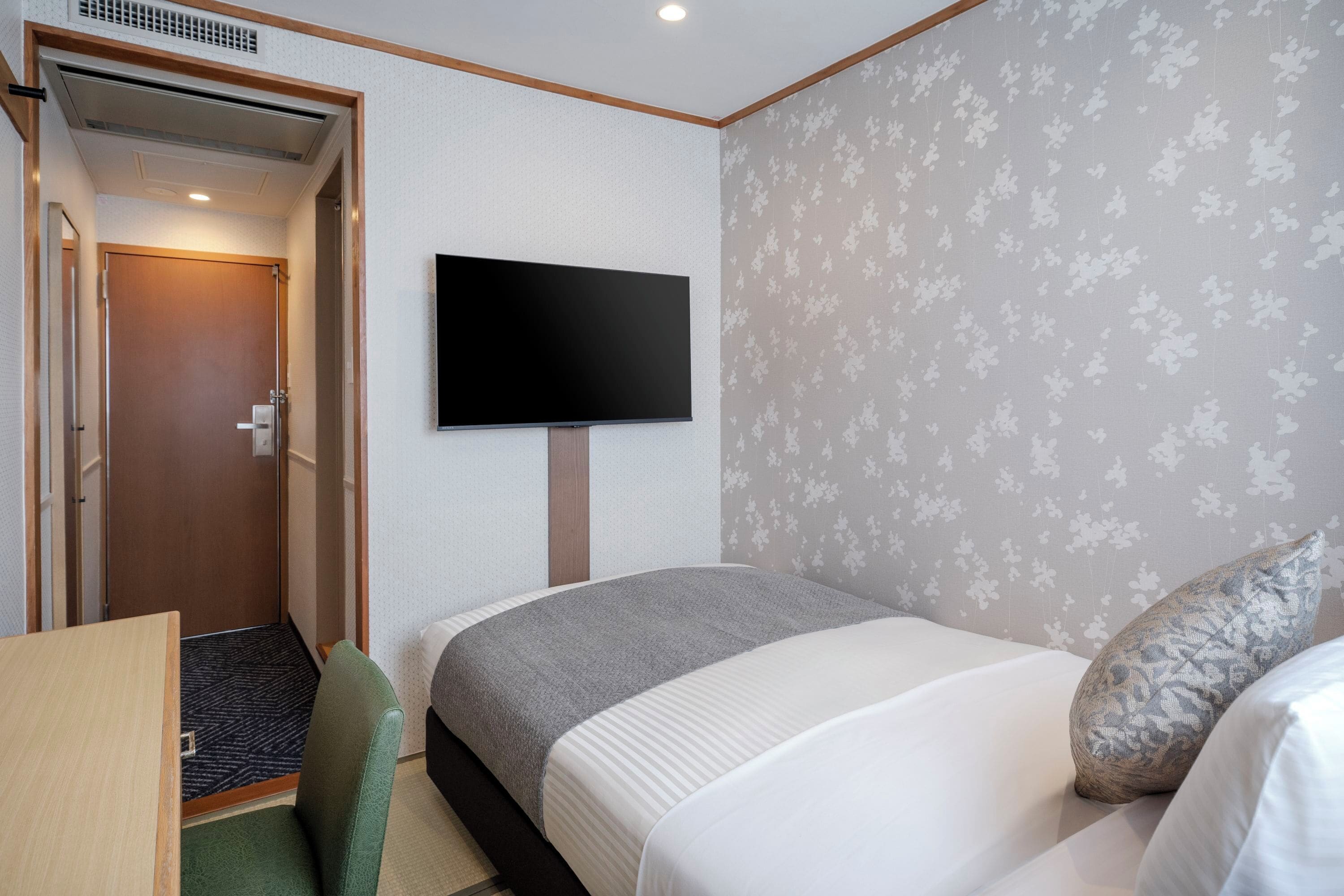Habitación de hotel con cama, televisión montada en la pared, una puerta y un pequeño escritorio con silla.