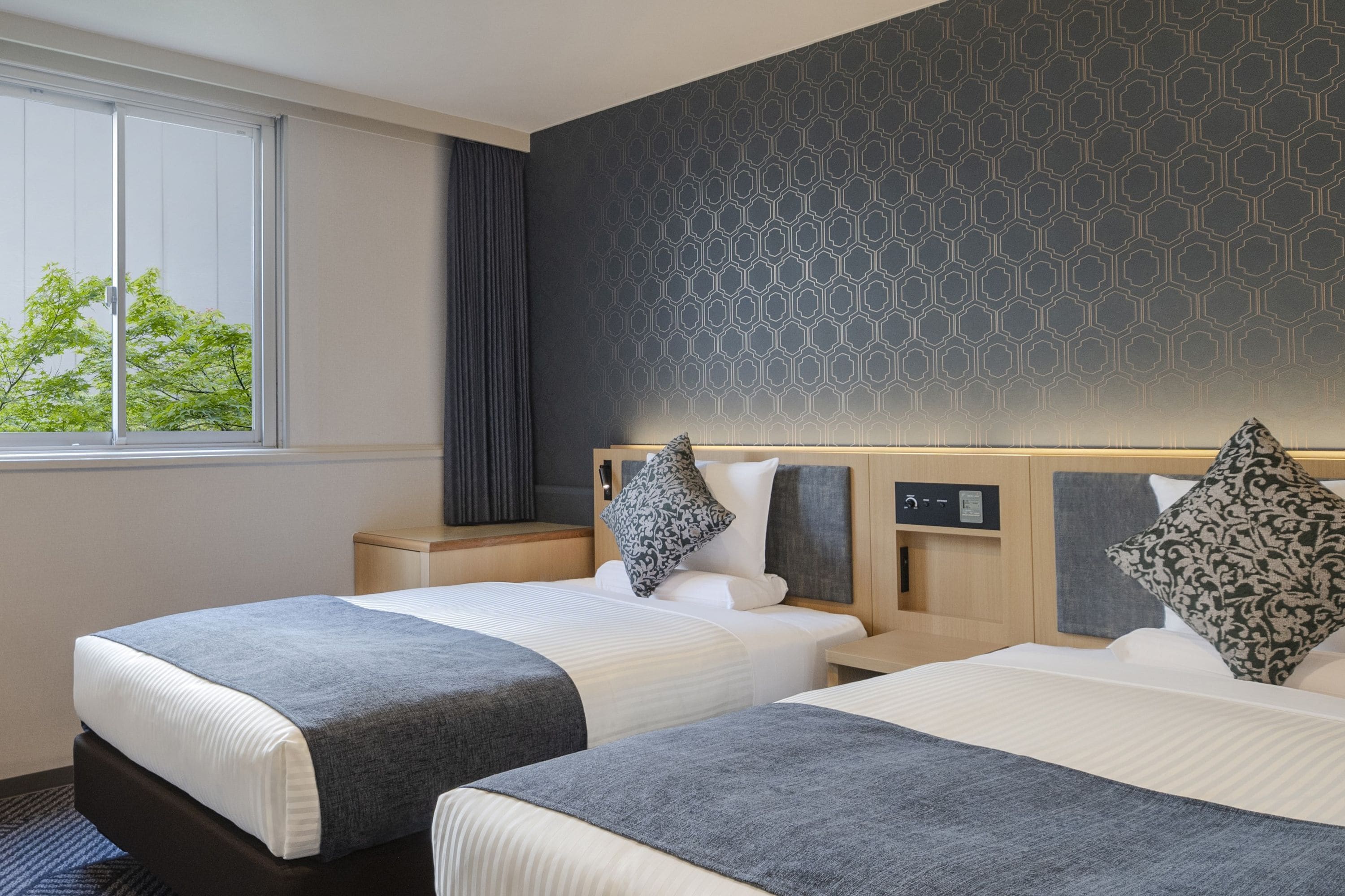 Une chambre d’hôtel avec deux lits, une fenêtre donnant sur la verdure, un mur décoré d’un motif et un panneau de commande sur la tête de lit.