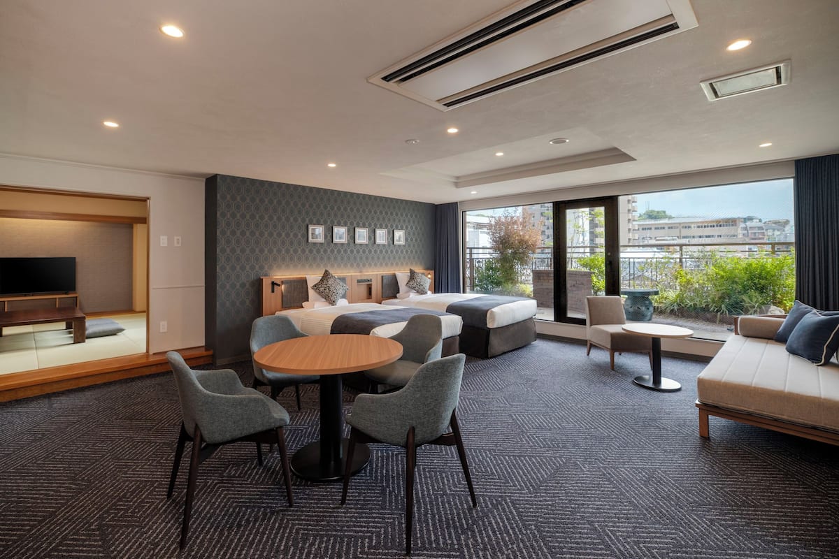 A modern hotel room with a sofa, a dining table, a TV, and a large window with a view.