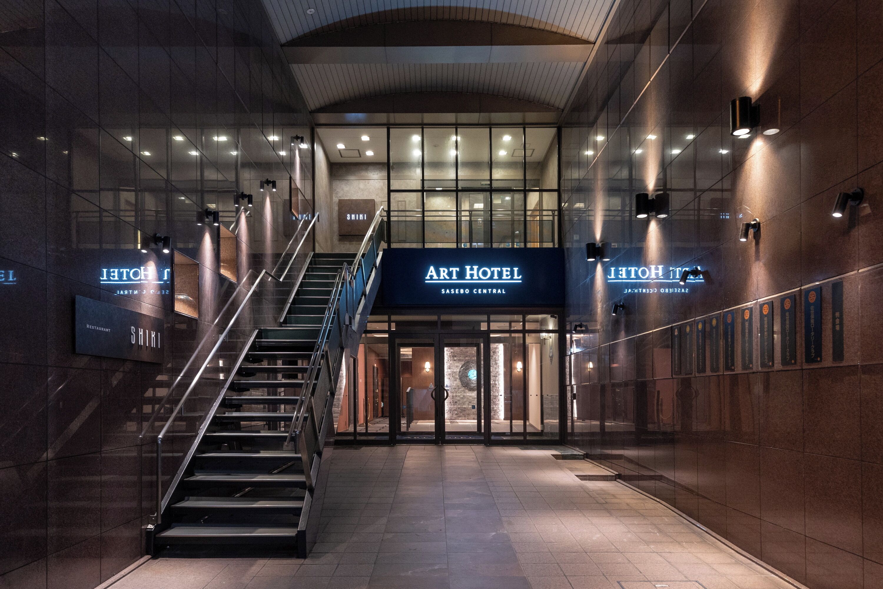 Uma entrada de hotel moderna com uma escadaria que leva ao saguão, uma placa indicando o Art Hotel e uma área de recepção.