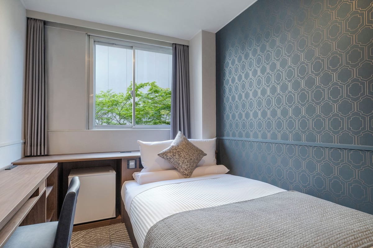 A hotel room with a bed, a desk, a chair, a small refrigerator, and a window with curtains.