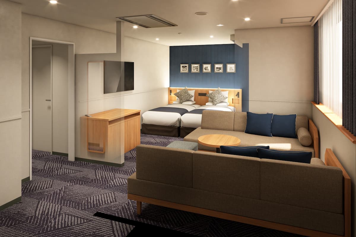 A modern hotel room with a sofa, a small table, a bed with a headboard, a wall-mounted TV, and a window with curtains.