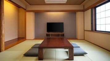 Una habitación con suelo de tatami, una mesa de madera, cojines, un televisor sobre un mueble y una ventana.