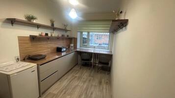 A compact kitchen with a refrigerator, a microwave, and a sink. There is a window with blinds and a small table with chairs.