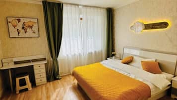 A bedroom with a bed, a desk, a chair, a nightstand, and a wall-mounted lamp.