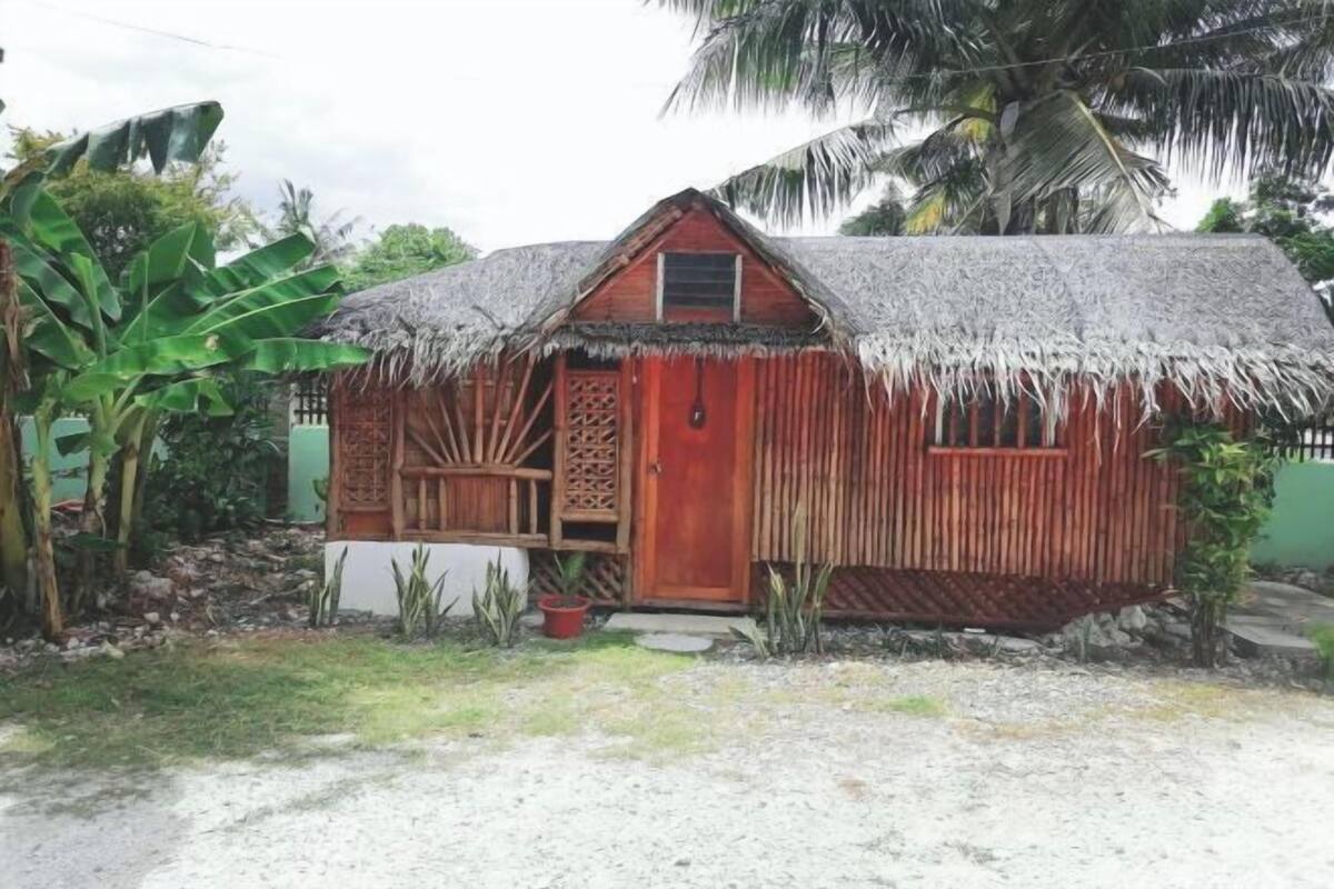 A small wooden house with a thatched roof, surrounded by tropical vegetation.
