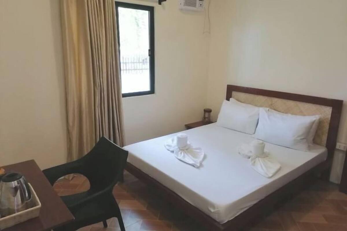 A hotel room with a bed, a chair, a table with a coffee pot, and a window with curtains.