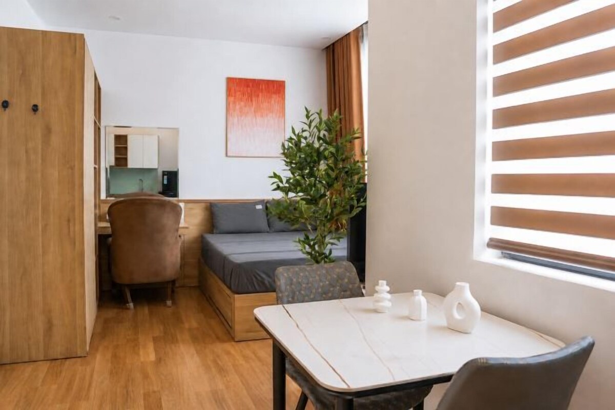 A compact room with a bed, a desk, a chair, a small table, and a window with blinds.