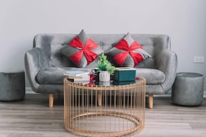 A grey sofa with red and grey pillows, a round wicker coffee table, and two grey ottomans. - Merci Vinhomes Marina Hai Phong (Haiphong)