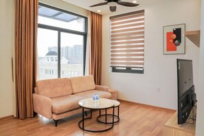 A room with a sofa, a round coffee table, a ceiling fan, a window with blinds, and a television. - Merci Vinhomes Marina Hai Phong (Haiphong)