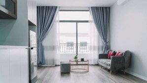 A modern living room with a large window, a round coffee table, a grey sofa, and a small round ottoman. - Merci Vinhomes Marina Hai Phong (Haiphong)