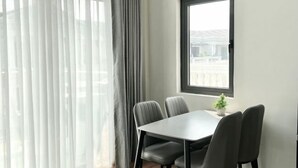 A dining area with a table and chairs, a window with a view, and a curtain. - Merci Vinhomes Marina Hai Phong (Haiphong)