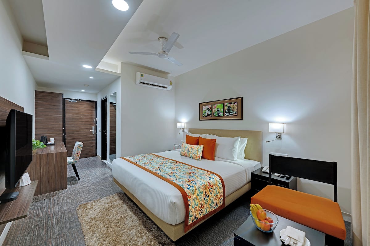 A hotel room with a bed, bedside tables, a desk, a chair, a TV, and an air conditioning unit.