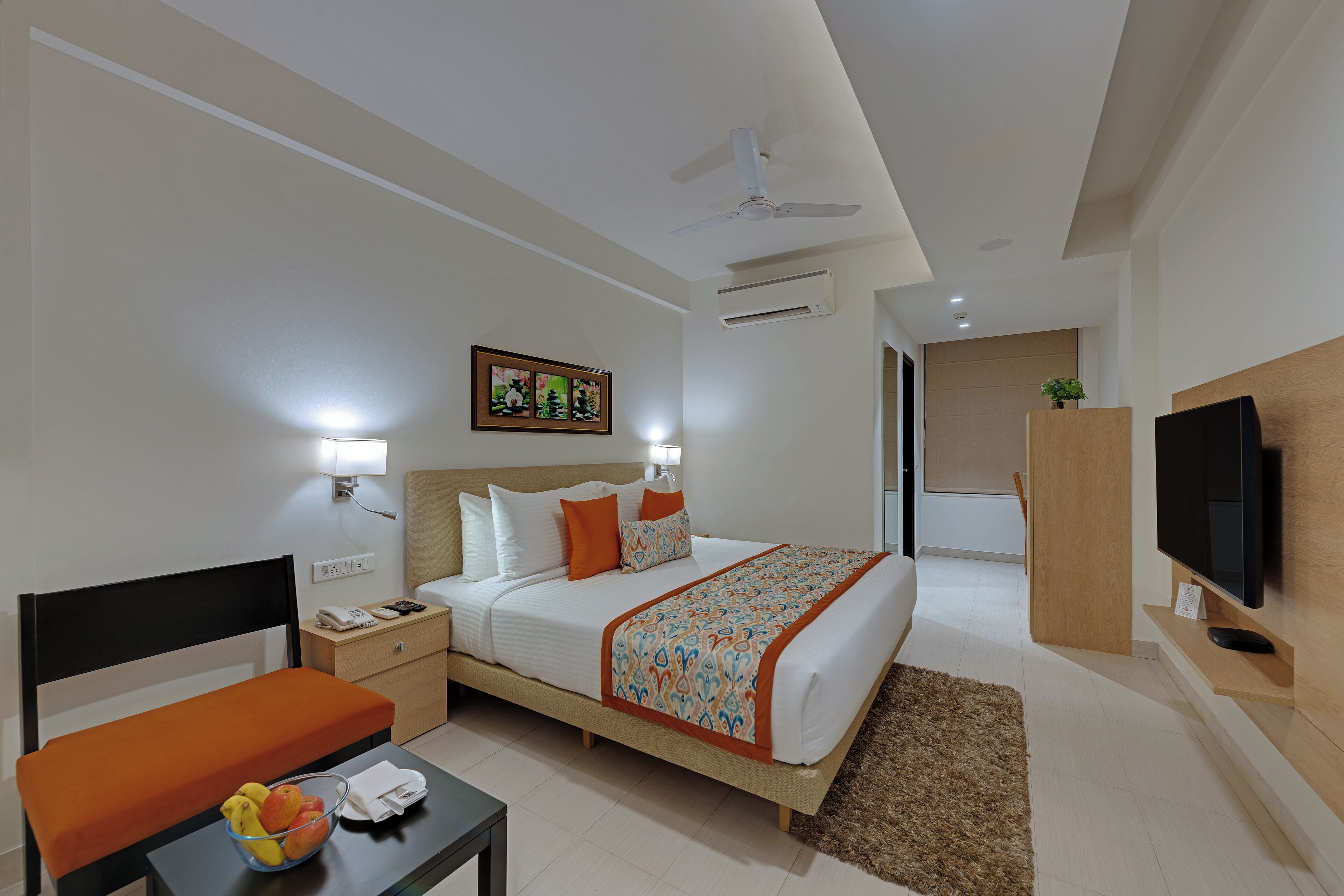 A modern hotel room with a bed, bedside table, chair, TV, and a small desk with a bowl of fruit.