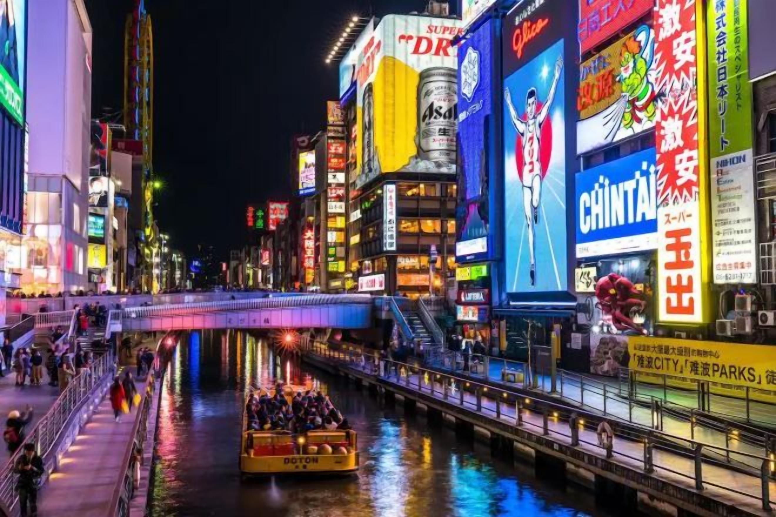 A brightly lit urban street with a river, illuminated billboards, and a pedestrian bridge.