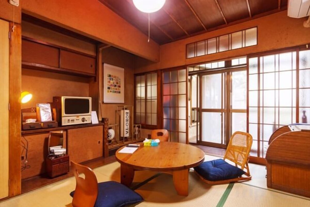 Una habitación japonesa tradicional con suelo de tatami, una mesa baja de tatami, un armario de madera bajo, un televisor y una puerta corredera.