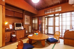 Una habitación japonesa tradicional con suelo de tatami, una mesa baja de tatami, un armario de madera bajo, un televisor y una puerta corredera.