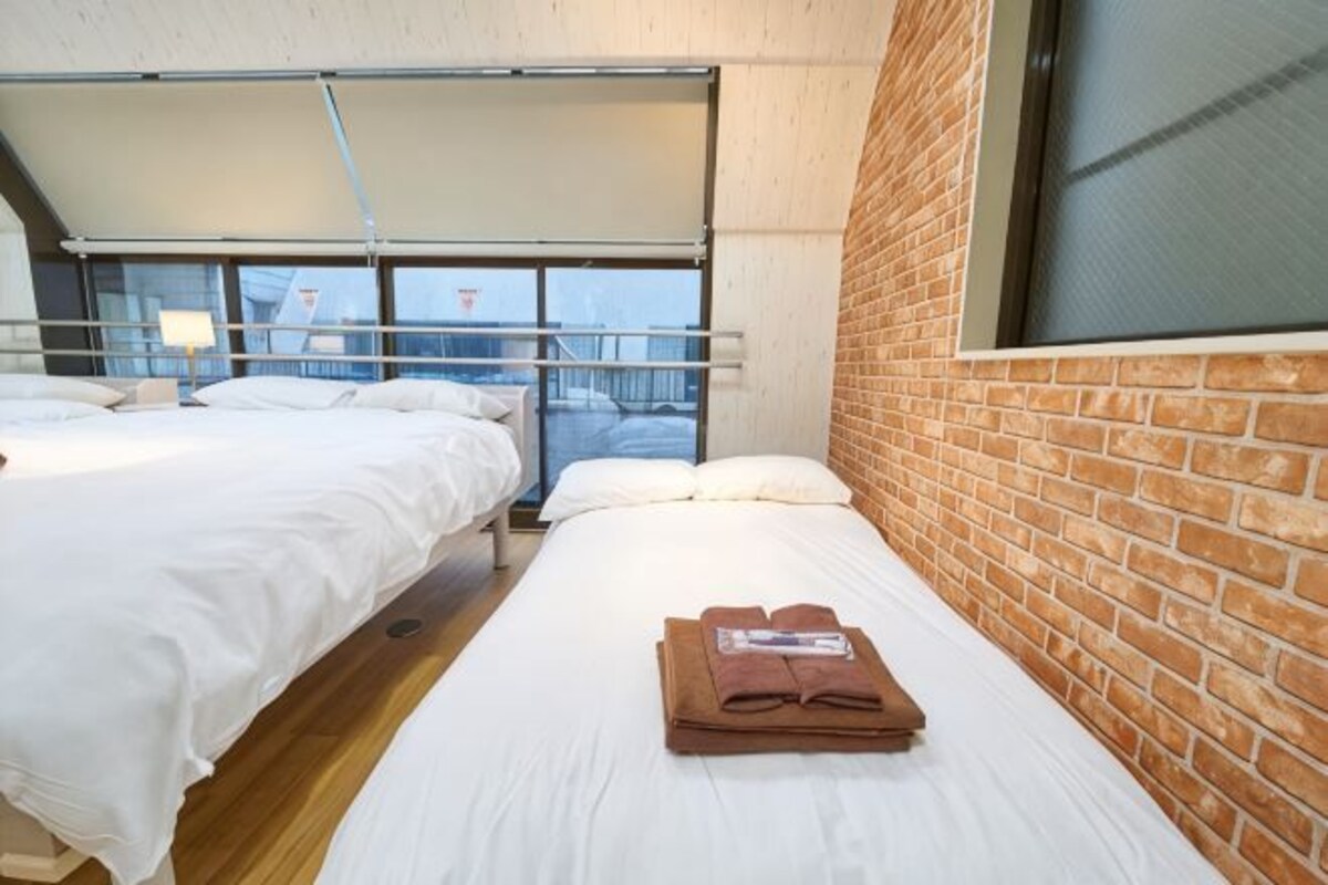 A room with two beds, a brick wall, and a window overlooking a balcony.