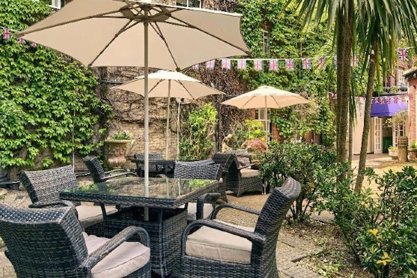 An outdoor patio with wicker furniture and umbrellas, surrounded by lush greenery and a stone wall.