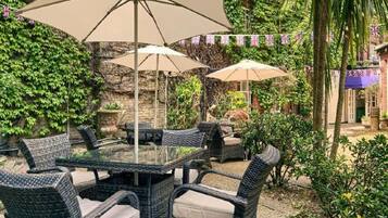 An outdoor patio with wicker furniture and umbrellas, surrounded by greenery and a stone wall.