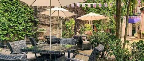 An outdoor patio with wicker furniture and umbrellas, surrounded by greenery and a stone wall.