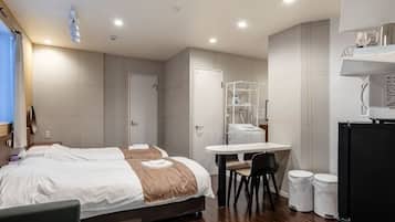 A hotel room with a bed, a desk, a chair, a refrigerator, and a trash bin.
