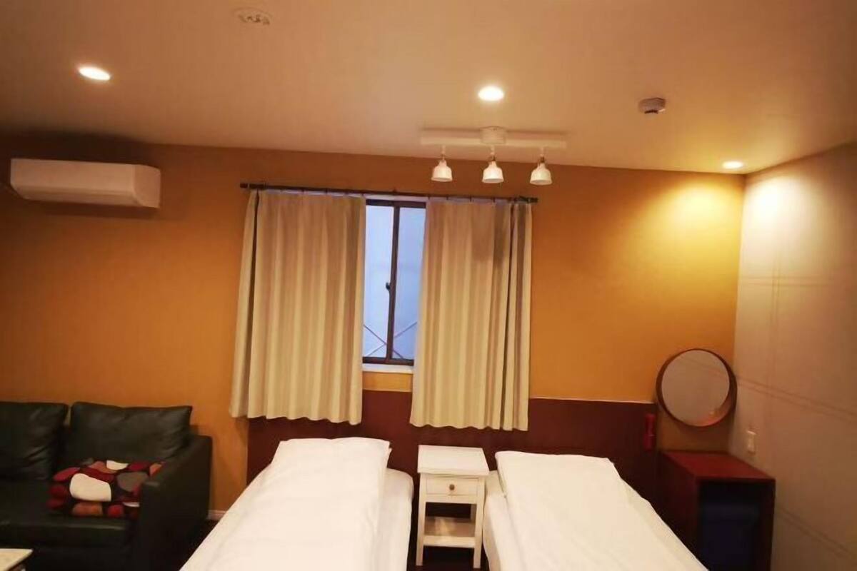 Une chambre d’hôtel avec deux lits, une petite table, une chaise, une fenêtre avec des rideaux, un appareil de climatisation et un miroir rond.