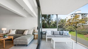 A modern living room with a grey sofa, a white coffee table, and a grey sectional sofa. There is a glass door leading to an outdoor balcony with a view of trees and buildings.
