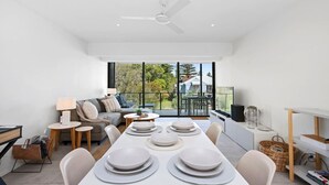 A modern living room with a dining area, large windows, and a balcony with a view of trees and houses.