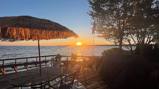 A wooden deck with a table and chairs, overlooking a body of water at sunset.