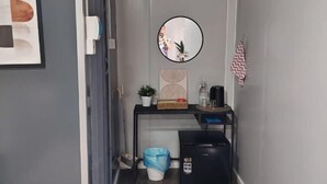 A compact room with a desk, a trash bin, a black printer, a mirror, and a framed artwork on the wall.