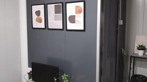 A room with a dark wall, a flat-screen TV, a potted plant, and three framed artworks.